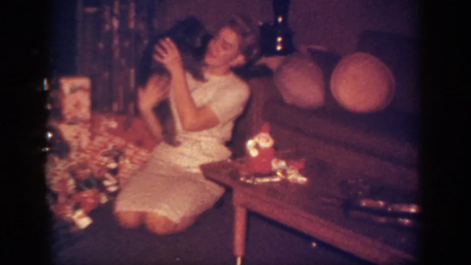 The woman embraces the puppy near a wooden coffee table with a Santa decoration in the 1962 Christmas home-movie scene.