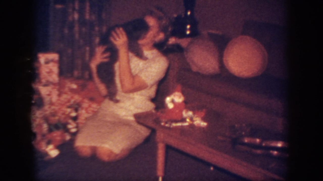 In a 1962 Blainsport, Pennsylvania home movie frame, a woman sits beside wrapped gifts and cuddles a small dark dog.