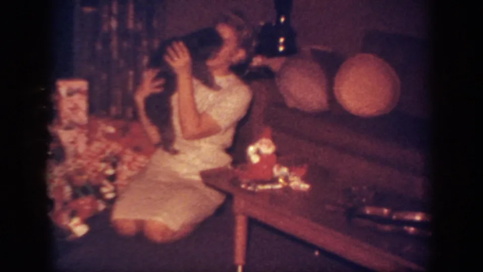 In a 1962 Blainsport, Pennsylvania home movie frame, a woman sits beside wrapped gifts and cuddles a small dark dog.