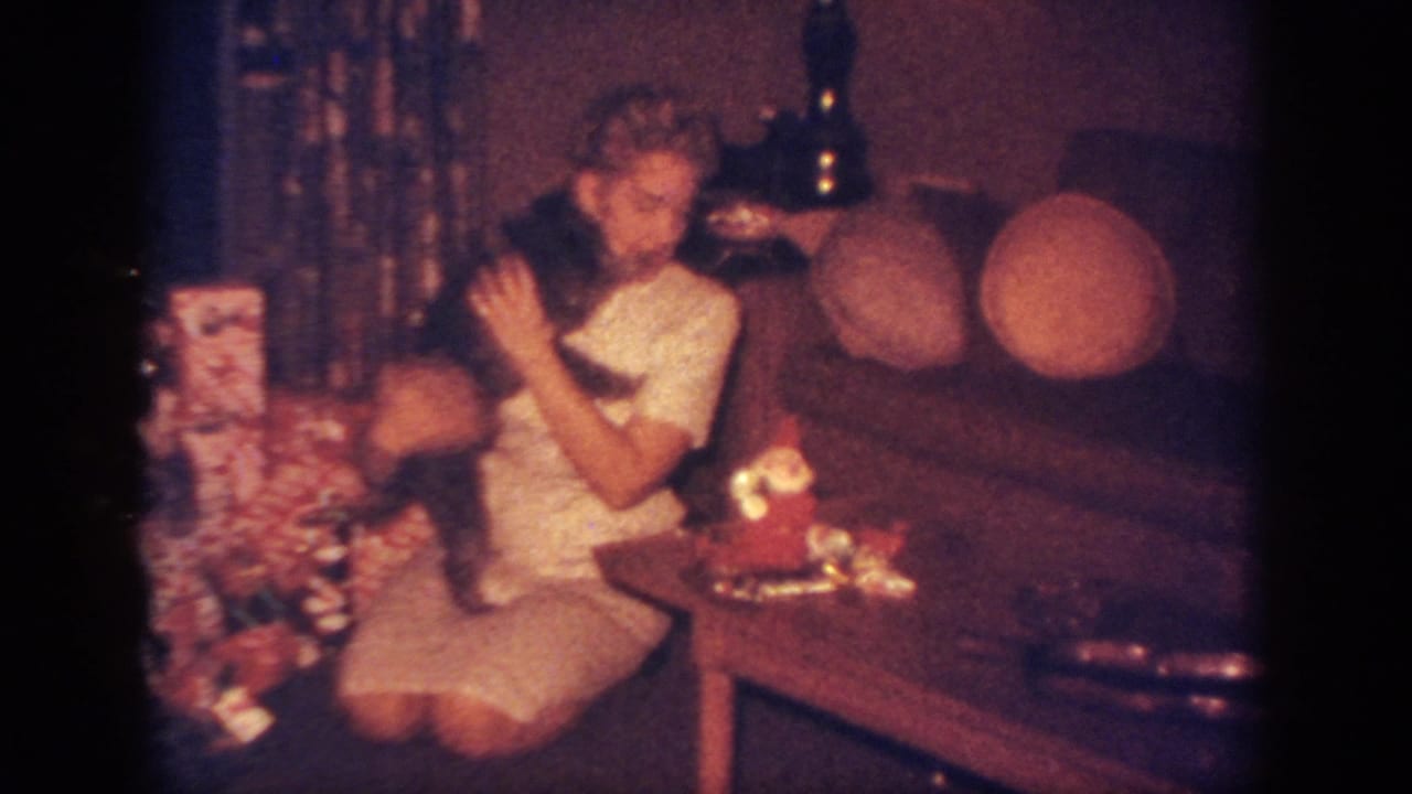 Woman kneels by wrapped Christmas gifts while holding a small dark dog against her shoulder.
