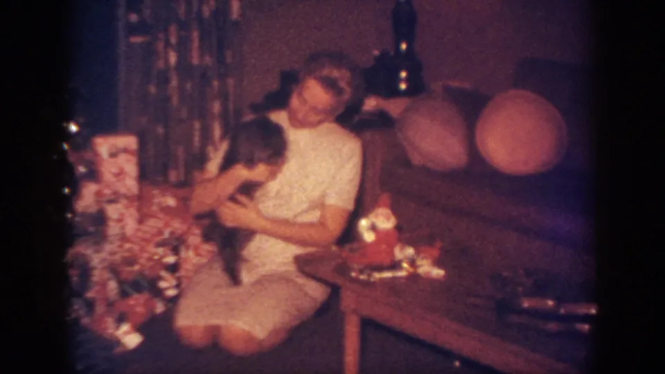 The home-movie frame shows the woman looking down at the dog near a tinsel Christmas tree and stacked gifts.