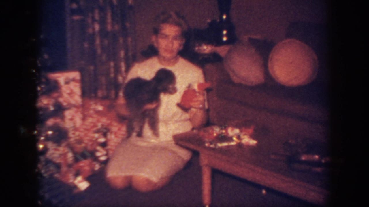 1962 Blainsport home-movie frame showing a woman in a white dress holding a small dark dog near a Christmas tree and gifts.