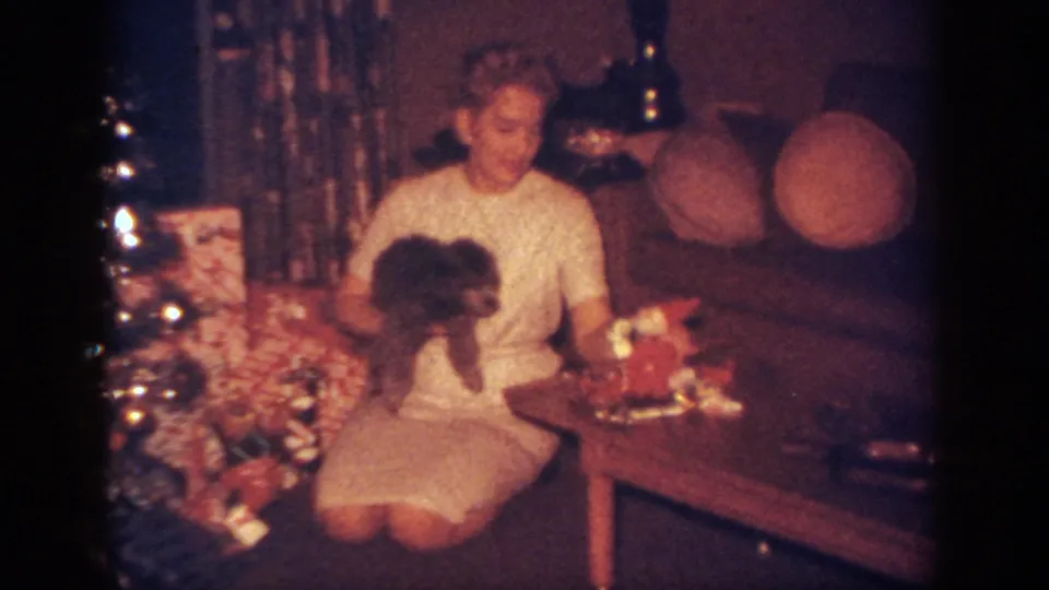 Blainsport 1962 holiday home-movie frame with a seated woman holding a dog amid presents, tree decorations, and living-room furniture.
