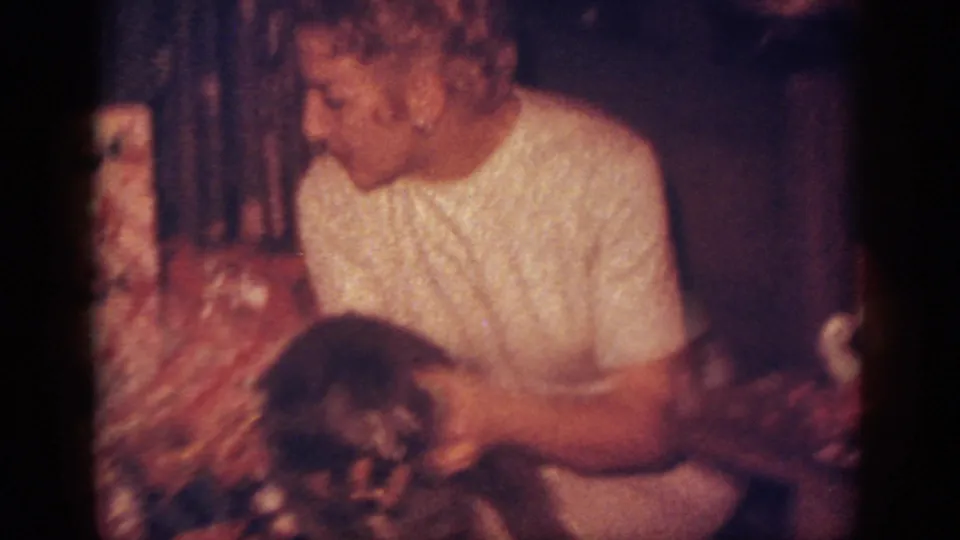 Close 1962 Blainsport frame of a woman in a white dress holding a small dark dog beside holiday decorations in a private living room.
