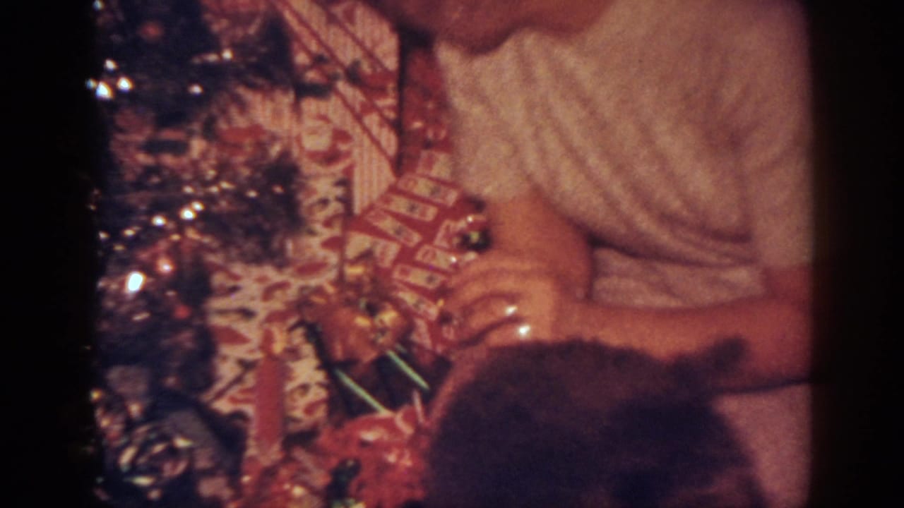 1962 Blainsport home-movie frame showing a woman beside a decorated Christmas tree with presents and a dark dog partly in view.