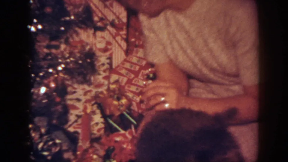 1962 Blainsport home-movie frame showing a woman beside a decorated Christmas tree with presents and a dark dog partly in view.