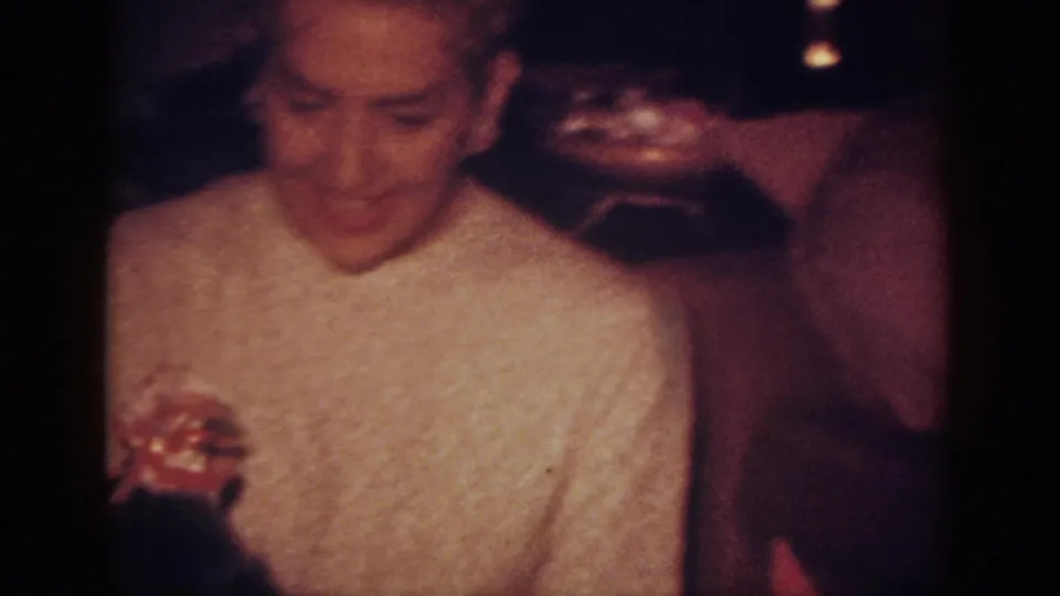 1962 Blainsport home-movie frame showing the woman in a light sweater looking toward a dark dog near her hands.