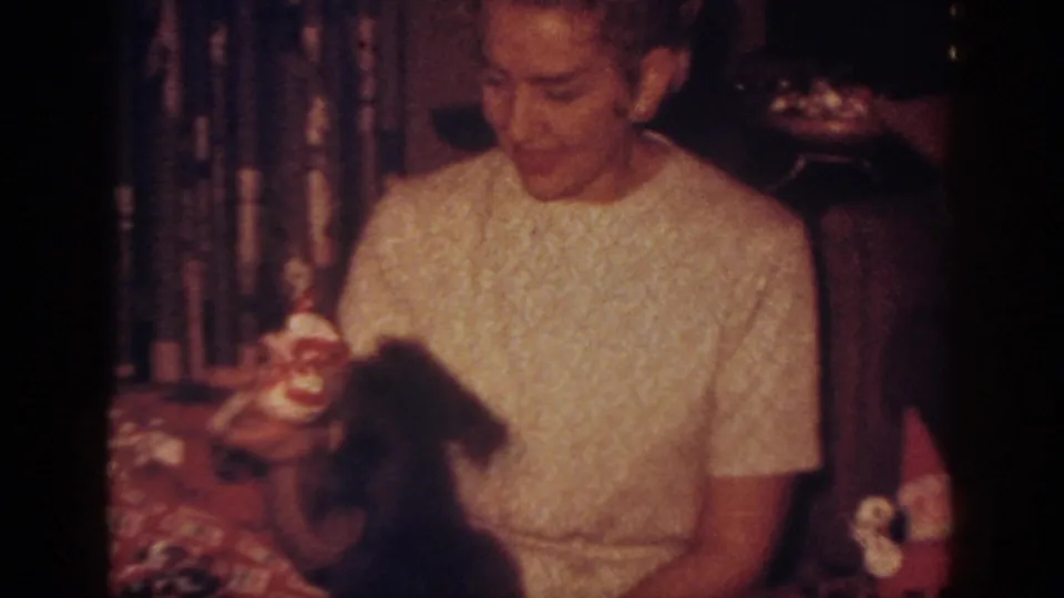 1962 Blainsport home-movie frame of a smiling woman playing with a dark dog beside Christmas gifts and decorations.