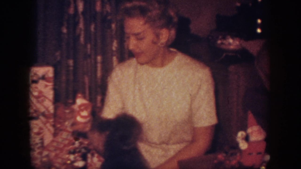 In a 1962 Blainsport, Pennsylvania home-movie frame, a woman sits near wrapped Christmas presents and a small dog beside the tree.