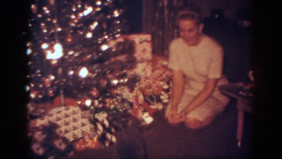 A woman kneels beside a bright Christmas tree and stacked presents as a small dark dog sits in the foreground.