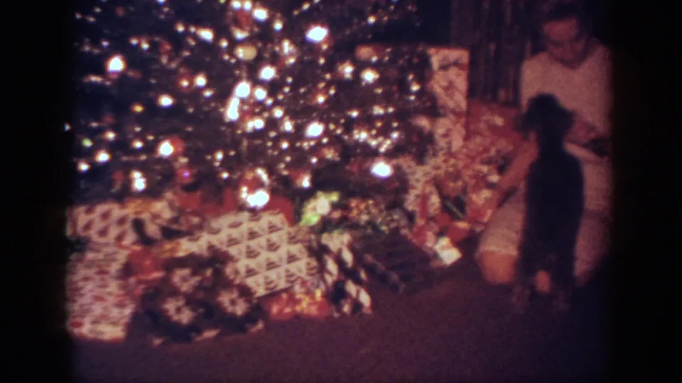 A small dark puppy stands near the seated woman at the edge of a brightly lit Christmas tree, with wrapped presents visible below.