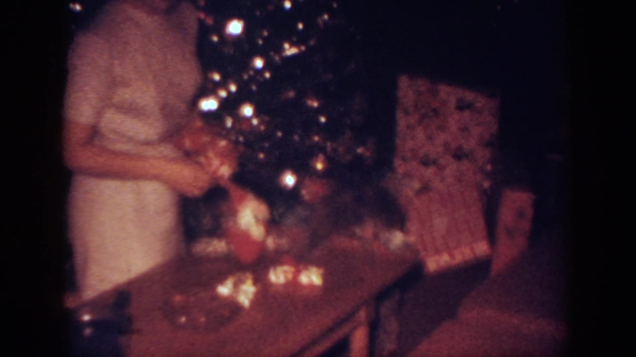 In a 1962 Blainsport home-movie frame, a woman in a pale dress stands by a glowing Christmas tree and wrapped gifts.