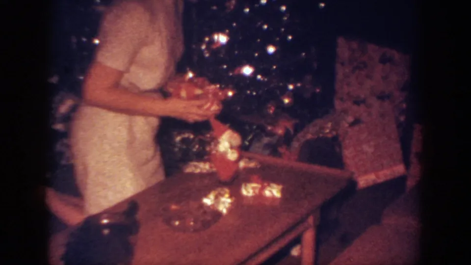 A close 8mm frame shows the woman near the decorated tree, with wrapped presents and a table in the living-room foreground.