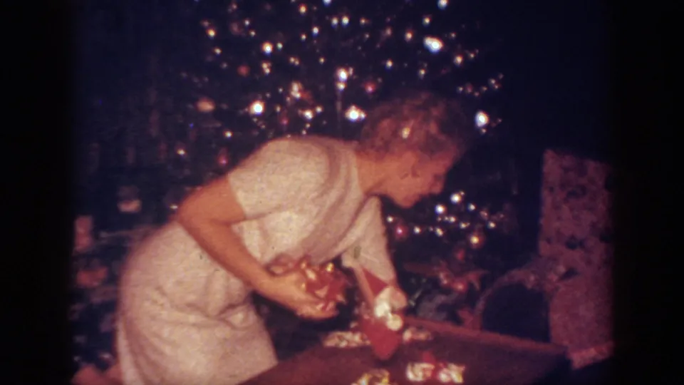 The woman bends toward the wrapped presents below the illuminated Christmas tree in the 1962 home interior.