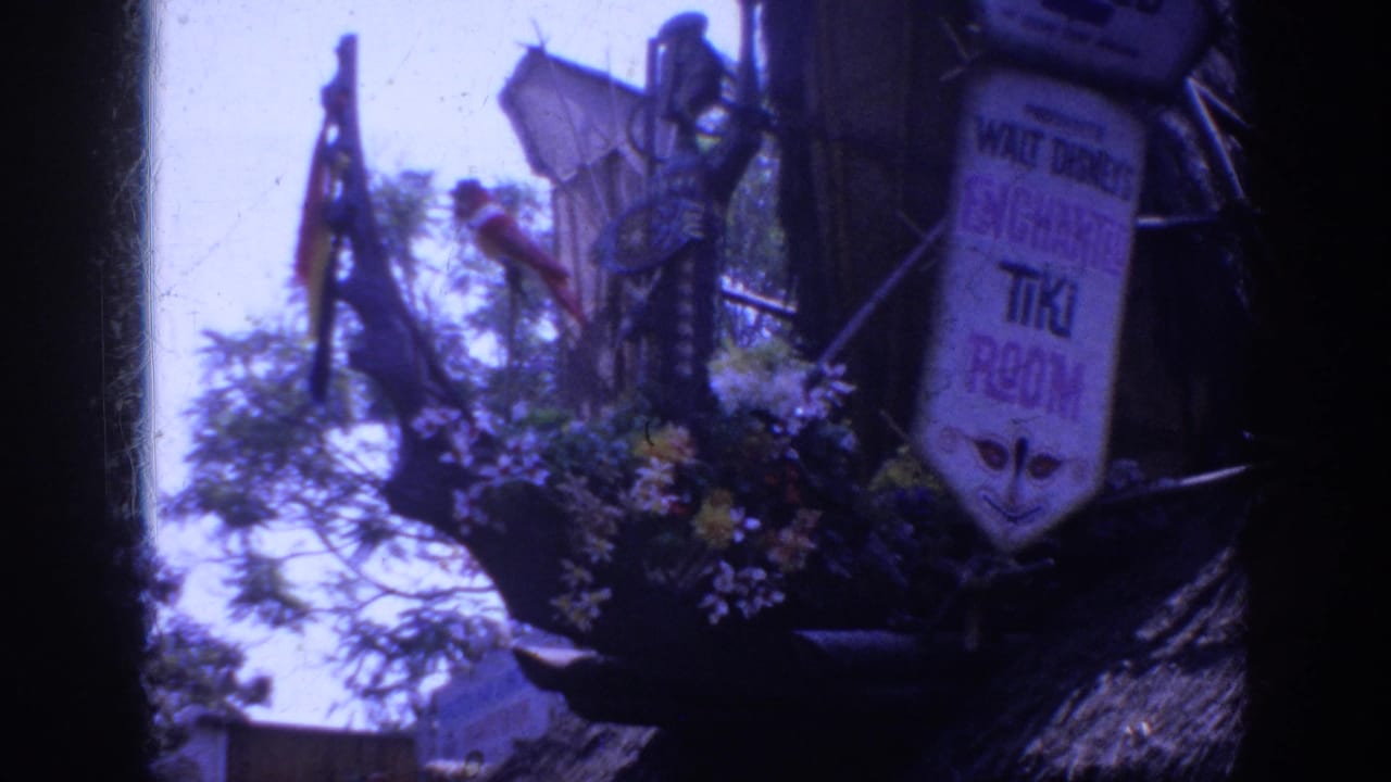 1962 Anaheim Disneyland home-movie frame showing the Enchanted Tiki Room entrance sign amid flowers, thatch, poles, and decorative bird forms.