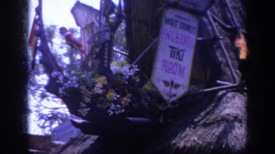 1962 Anaheim Disneyland frame of the Enchanted Tiki Room sign with flowers, thatch, branchwork, and a small flame torch at the right edge.