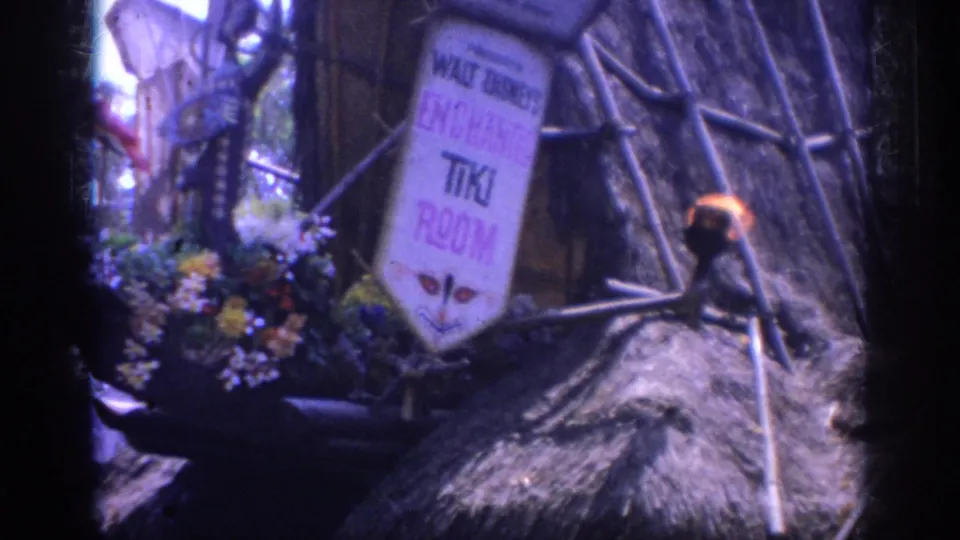 1962 Anaheim Disneyland home-movie still showing the Tiki Room sign near flowers, thatched roofing, pale poles, and a bright torch flame.