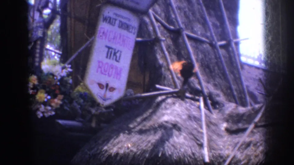 1962 Anaheim Disneyland frame holding on the Enchanted Tiki Room sign with flowers, thatch, crossed branches, and torchlight.