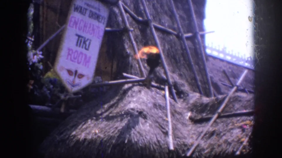 1962 Anaheim home-movie frame showing the Enchanted Tiki Room exterior sign, thatched roof, bamboo supports, and a burning torch.