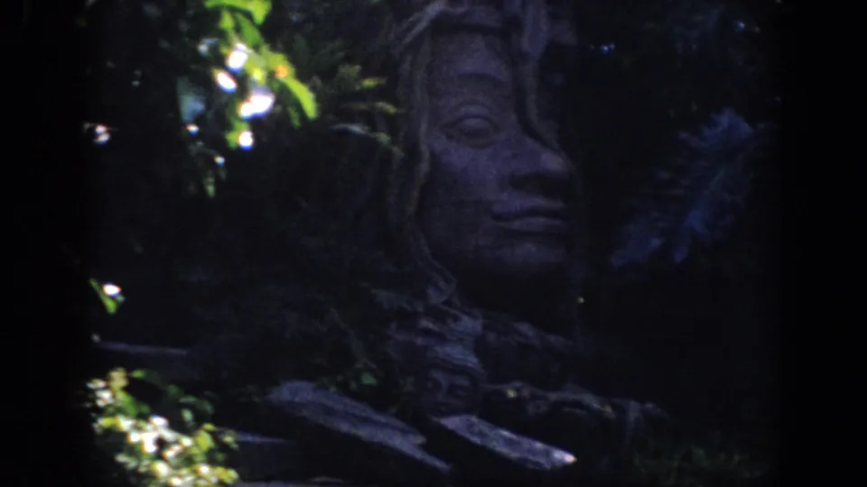 1962 Anaheim 8mm frame showing a carved stone face sculpture set among plants in low-light themed attraction scenery.