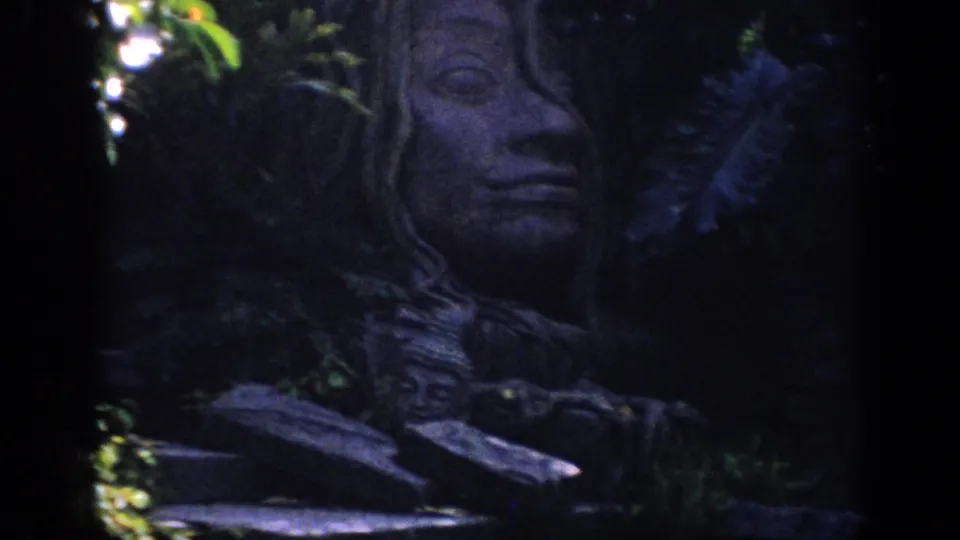 1962 Anaheim 8mm frame showing a large Buddha-style stone face emerging from shadowed garden foliage.