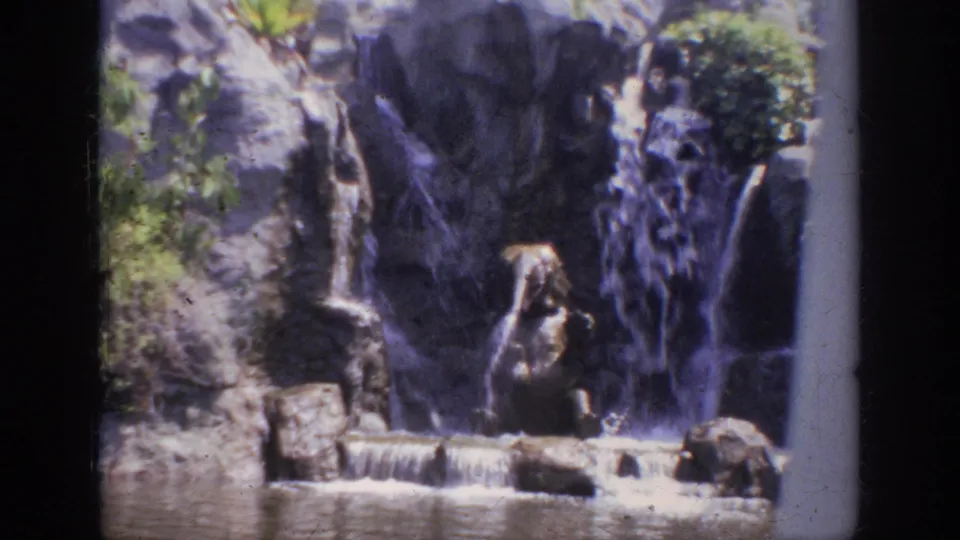 1962 Anaheim home-movie frame of multiple small waterfall streams over rockwork above lagoon water.