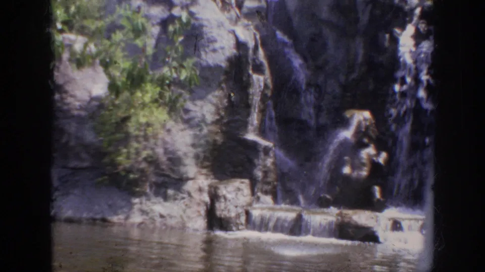 1962 Anaheim 8mm view of a landscaped rock waterfall with greenery and rippling lagoon water.