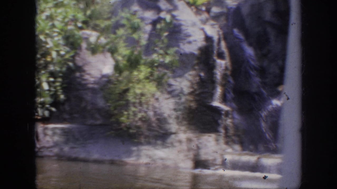 1962 Anaheim home-movie frame showing a rocky waterfall feature beside shallow water, with soft color and dark 8mm frame edges.
