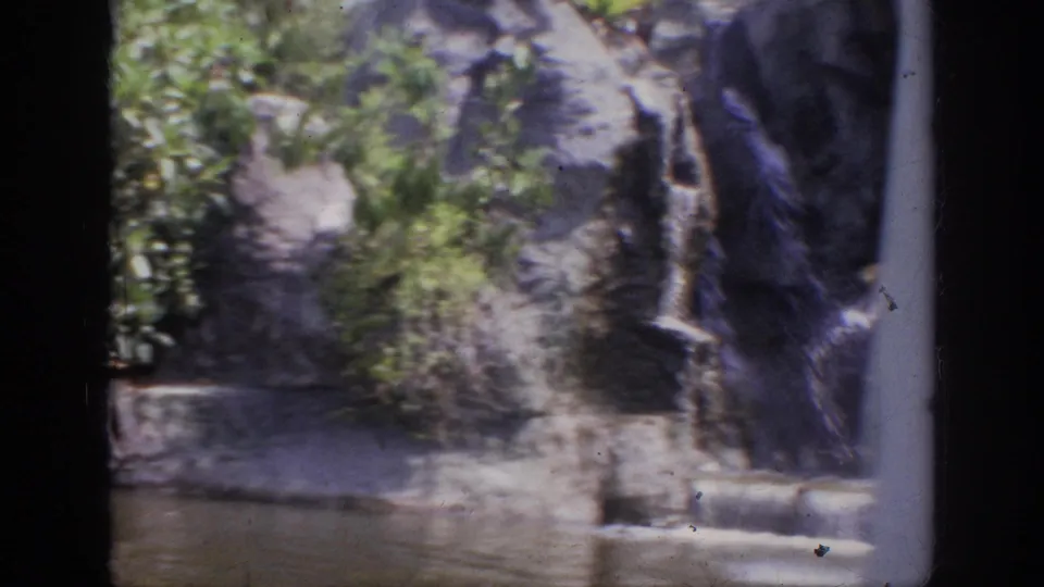 1962 Anaheim home-movie frame showing a rocky waterfall feature beside shallow water, with soft color and dark 8mm frame edges.