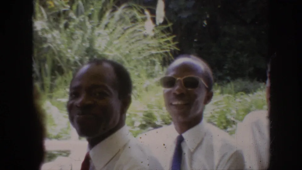 Final 1962 Anaheim frame showing two smiling adult men in white shirts and ties, with greenery behind them and part of another person at the edge.