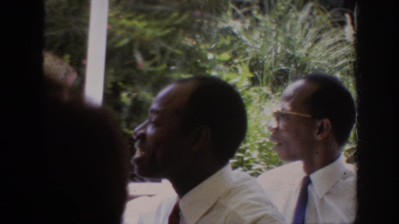 Several Black adult men in white shirts and ties converse outdoors near greenery.