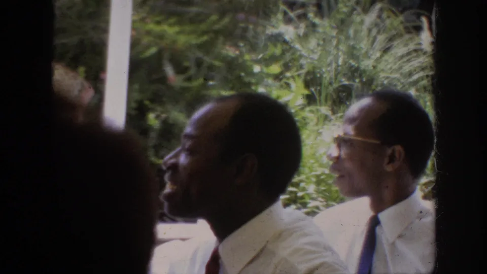1962 Anaheim home-movie frame with Black adult men in dress shirts and ties talking beside dense foliage and a pale frame.