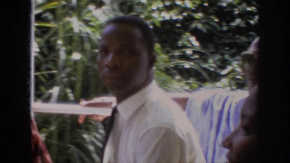 1962 Anaheim 8mm frame showing a Black adult man in a white dress shirt facing the camera near foliage and partially visible companions.