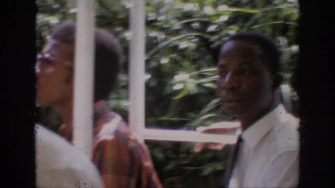 1962 Anaheim home-movie frame showing adult male riders behind white ride railings with leafy foliage in the background.