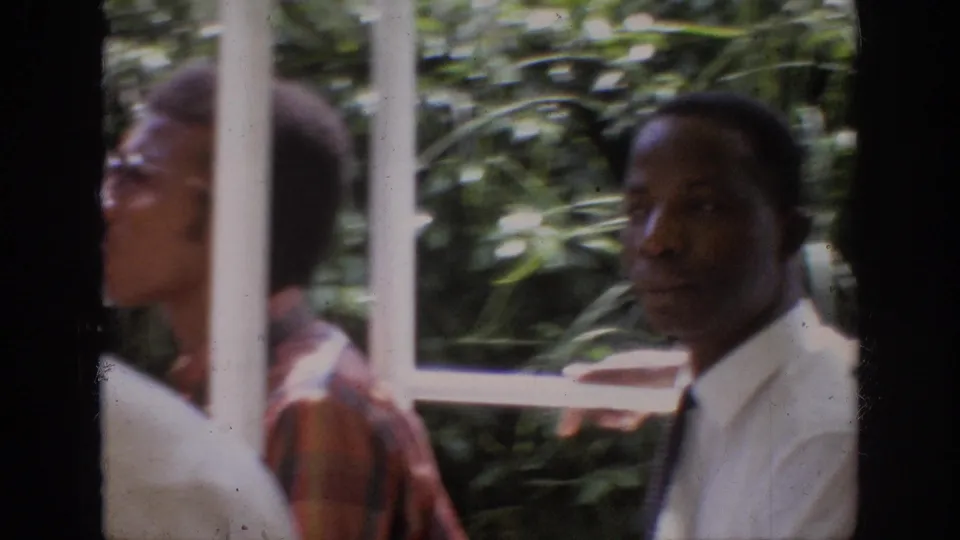 1962 Anaheim home-movie frame showing adult male riders behind white ride railings with leafy foliage in the background.