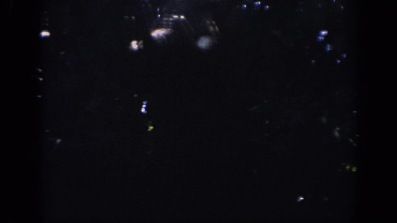 Mostly dark frame with small points of light and film grain.