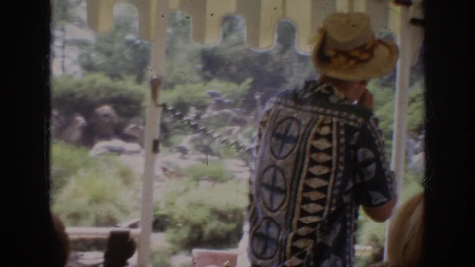A passenger in patterned clothing stands near a vehicle post as the 1962 Anaheim footage looks toward rocks, plants, and animal figures.