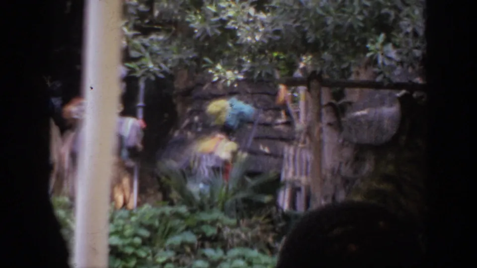 1962 Anaheim home-movie frame shows costumed performers with bright headdresses near plants and a rustic themed structure, partly blocked by foreground framing.