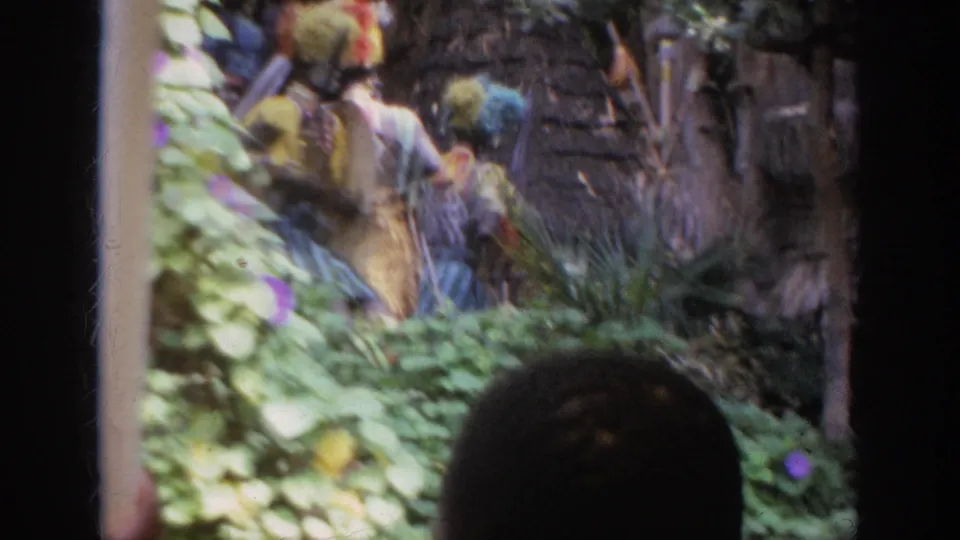 1962 Anaheim visitor-shot footage shows costumed performers in colorful headdresses beside a rustic wall and planted area, seen past a foreground silhouette.