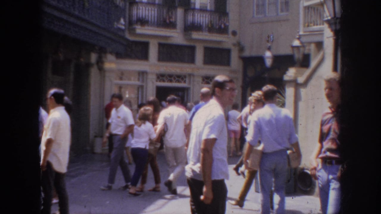 1962 Anaheim home-movie frame showing pedestrians in a sunlit outdoor shopping district with storefronts and upper balconies.
