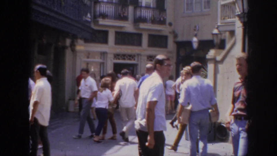 1962 Anaheim home-movie frame showing pedestrians in a sunlit outdoor shopping district with storefronts and upper balconies.