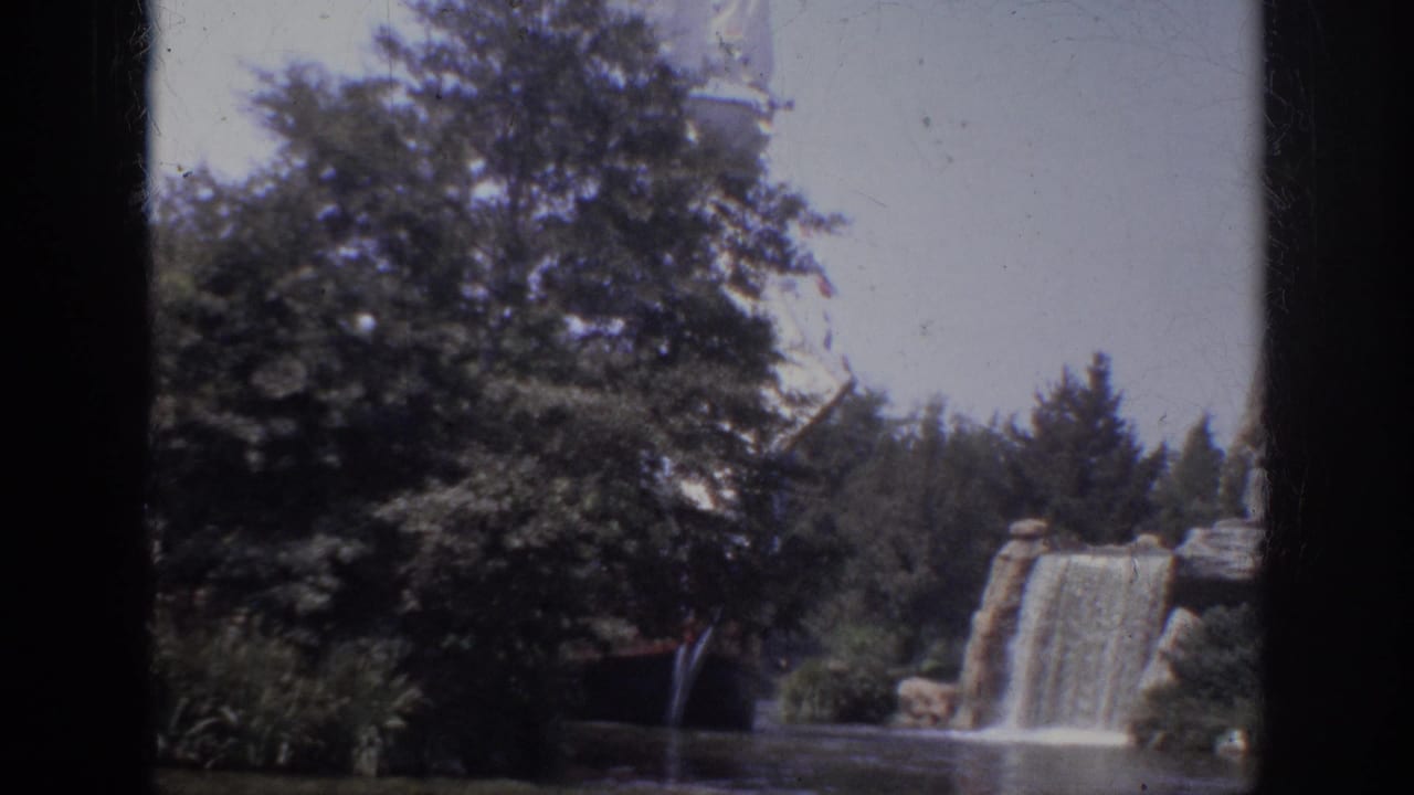 1962 Anaheim 8mm frame showing a landscaped lagoon, leafy trees, a rock waterfall, and a large white sail partly obscured by foliage.