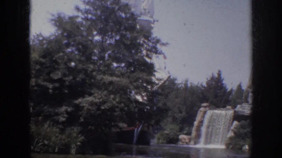 1962 Anaheim 8mm frame showing a landscaped lagoon, leafy trees, a rock waterfall, and a large white sail partly obscured by foliage.