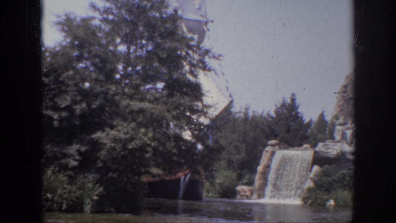Tree-lined lagoon with a white sail behind foliage and a waterfall flowing over rockwork.