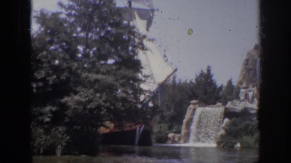 1962 Anaheim 8mm frame of a large white sail moving behind trees near a landscaped rock waterfall and lagoon.