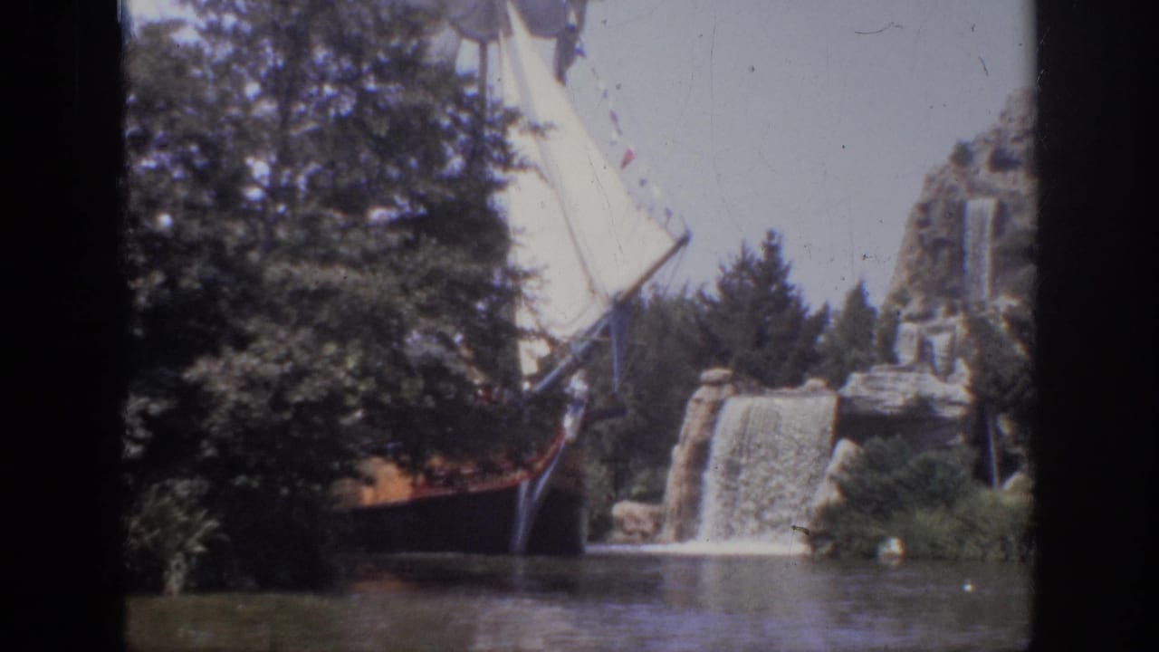 1962 Anaheim 8mm frame of a themed sailing ship passing landscaped rocks and a small waterfall beside a river channel.