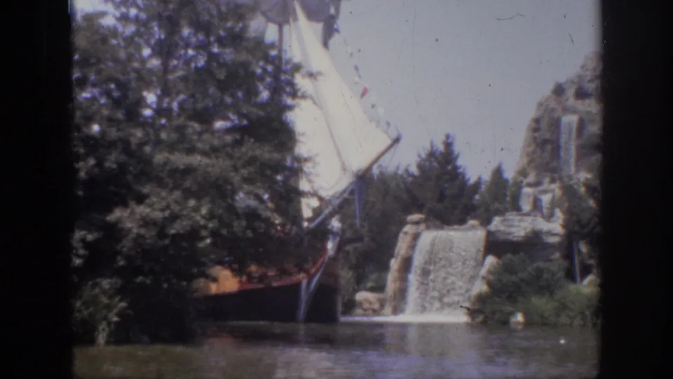 1962 Anaheim 8mm frame of a themed sailing ship passing landscaped rocks and a small waterfall beside a river channel.