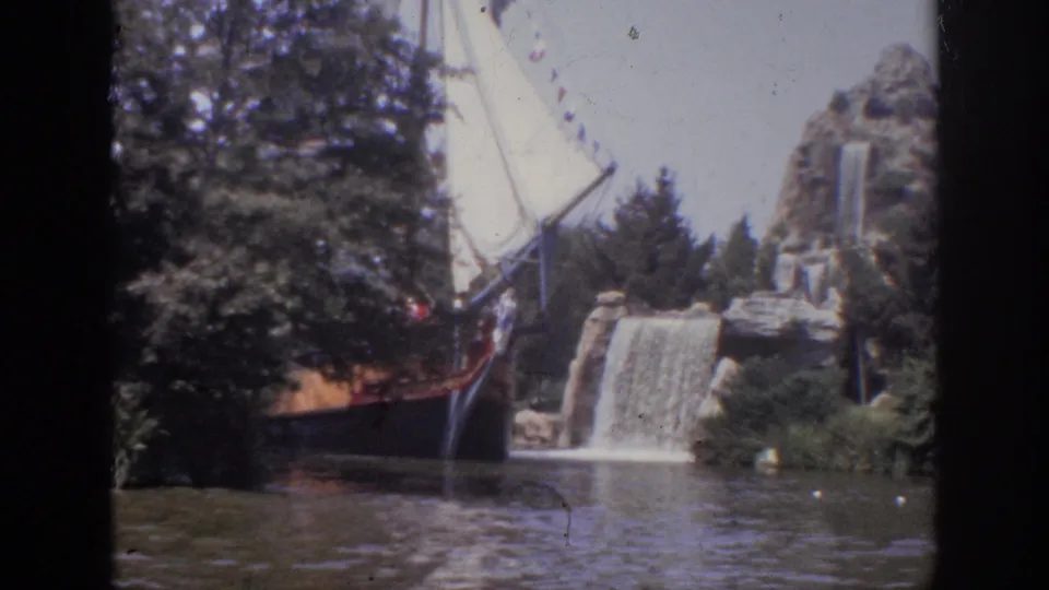 Visitor-shot 1962 Anaheim frame with the boat nearer the foreground, white sail above trees, and a cascading waterfall behind rockwork.