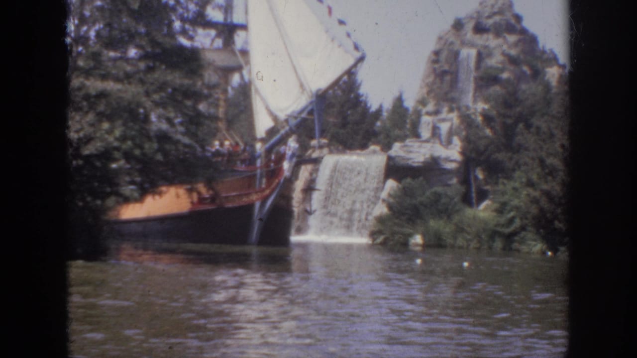 1962 Anaheim visitor-shot frame with a sail-rigged ride boat, artificial rockwork, waterfall, trees, and rippled water.