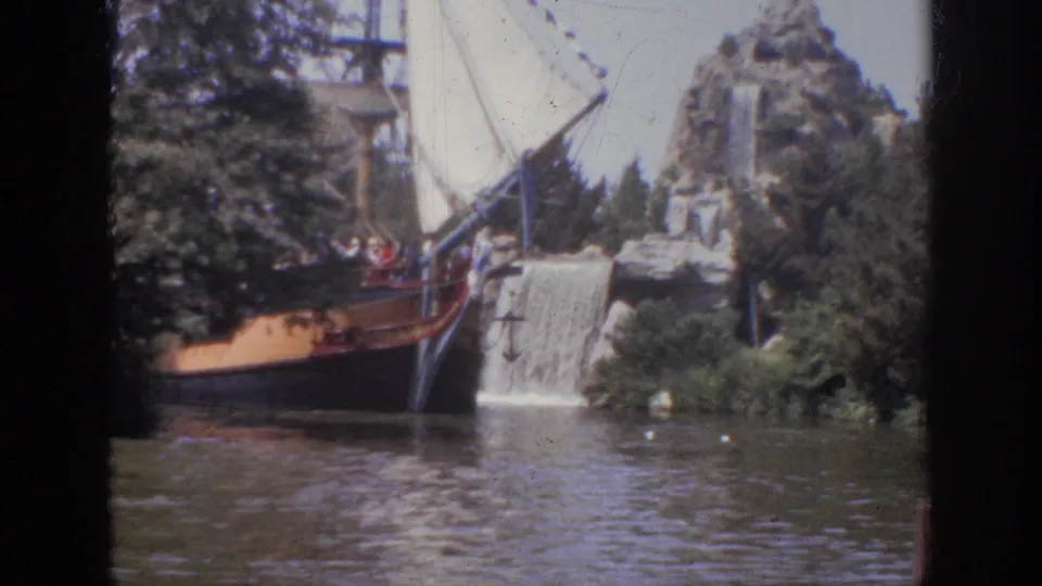 1962 Anaheim 8mm frame showing a wooden attraction boat crossing the lagoon in front of a rocky waterfall scene.